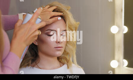 Stilvolle blonde Junge im Beauty Salon. Mädchen macht einen Abend Frisur am professionellen Friseur. Haarstilist verwendet Spray-förmige Haarspray cur zu beheben Stockfoto