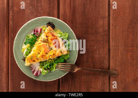 Ein Stück französische Quiche mit Lachs, mit grünem Salat Blätter, Schuß von der Oberseite in einem dunklen Holzmöbeln im Landhausstil Hintergrund mit Kopie Raum Stockfoto