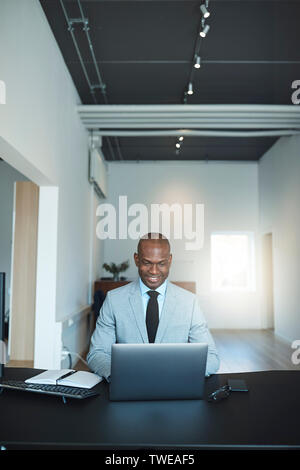 Lächelnden jungen afrikanischen amerikanischen Geschäftsmann online arbeiten mit einem Laptop an seinem Schreibtisch in einem modernen Büro Stockfoto