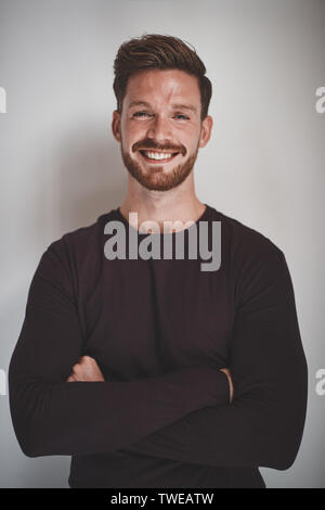 Beiläufig junge männliche Unternehmer Lächeln, während er mit seinen Armen vor grauem Hintergrund gekreuzt gekleidet Stockfoto