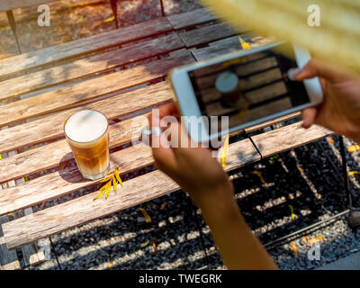 Woman's Hände halten weiß Smartphone und ein Foto von Iced Cappuccino in Glas auf Holztisch, Ansicht von oben Stockfoto