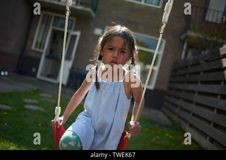 Nette junge asiatische Mädchen mit einem Kleid spielen auf einer Schaukel im Garten hinter dem Haus im hellen Sonnenschein Stockfoto