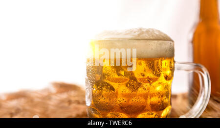 Krug Voller Bier mit Schaum und Flasche hinter auf Holztisch mit Gerste Ohren mit weißem Hintergrund eingerichtet. Horizontale Komposition. Vorderansicht. Stockfoto