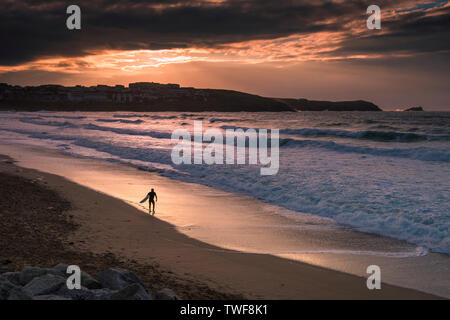 Einen spektakulären Sonnenuntergang als einsame Surfer sein Surfbrett zu Fuß, entlang der Küstenlinie von Fistral Beach in Newquay in Cornwall. Stockfoto