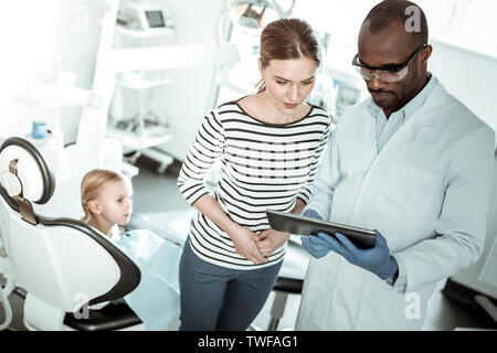 Zahnarzt Akzentuierung von Aufmerksamkeit. Executive stomatologist Holding Tablet und zeigen Ergebnisse der Beobachtung zu sorgen Mutter Stockfoto