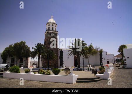 TEGUISE, LANZAROTE - juin 2013 3. 2019: Blick über Platz mit Palmen auf alten weißen Kirche gegen den blauen Himmel Stockfoto