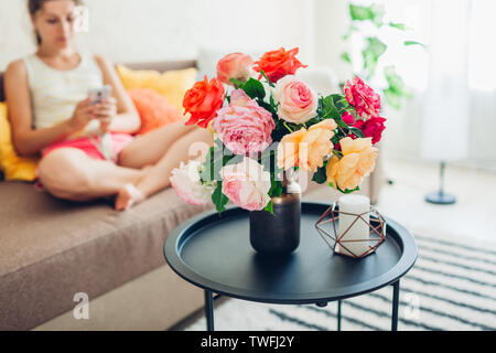 Glückliche junge Frau mit Smartphone sitzen auf der Couch zu Hause und entspannen. Wohnzimmer eingerichtet mit Blumenstrauß aus Rosen Stockfoto