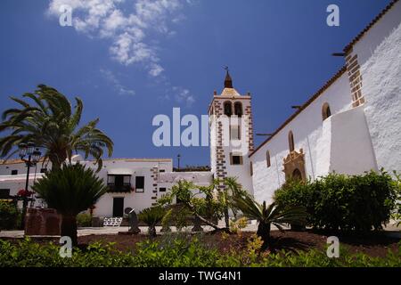 BETANCURIA, Fuerteventura - JUIN 14. 2019: Blick auf den Garten mit Palmen auf alten weißen Kirche mit Glockenturm gegen den blauen Himmel Stockfoto