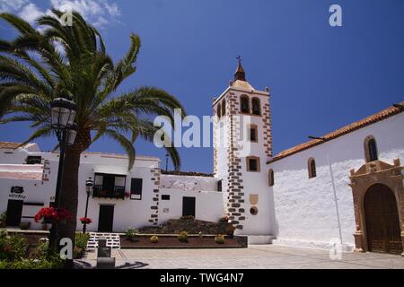 BETANCURIA, Fuerteventura - JUIN 14. 2019: Blick auf den Garten mit Palmen auf alten weißen Kirche mit Glockenturm gegen den blauen Himmel Stockfoto