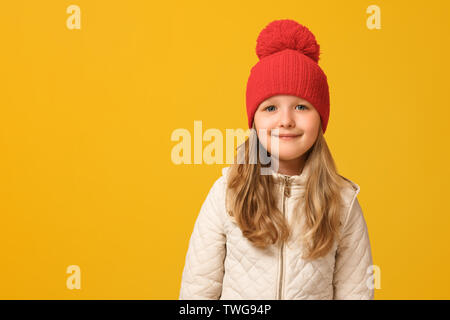 Porträt von einem fröhlichen Kind in eine beige Jacke und einen roten Hut auf einem gelben Hintergrund. Kleines Mädchen blond sieht in die Kamera. Herbst Konzept. Kopieren Sie sp Stockfoto