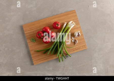 Tomaten, Frühlingszwiebeln und Knoblauch auf einer hölzernen Schneidebrett gegen eine moderne Beton Tischplatte Stockfoto
