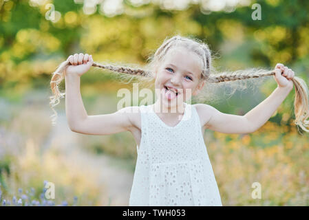 Funny Girl holding Haar Zöpfe. Kleine süße Mädchen mit langen blonden Haaren, die Zunge im Freien. Kid Emotionen zum Ausdruck zu bringen. April Stockfoto