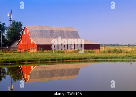 Eine klassische rote Scheune mit Windmühle im Teich im ländlichen Mittleren Westen an einem Sommertag mit blauem Himmel in Missouri, USA Stockfoto