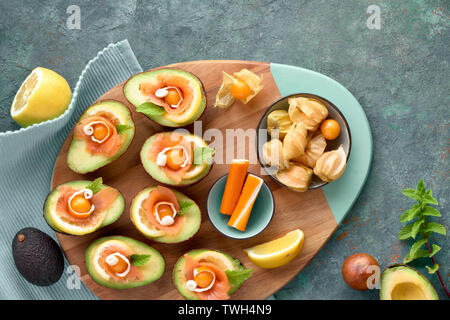 Avocado Boote mit Räucherlachs, Crab Sticks, Zitrone, Physalis und Minzeblatt, Ansicht von oben auf der cyanfarbenen strukturierten Hintergrund Stockfoto