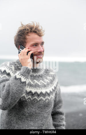 Smartphone Mann auf smart phone Wandern auf schwarzem Sand Strand auf Island isländische Pullover tragen durch den Ozean Meer. Stockfoto