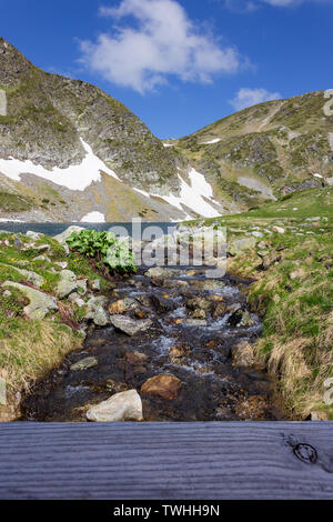 Schönen leuchtenden Farben eines Creek Streaming von berühmten Niere Lake im Rila Gebirge, durch hölzerne Brücke Zaun und tiefen blauen Himmel gerahmt Stockfoto