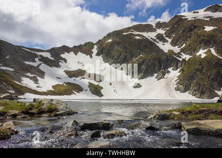 Low Angle View des Auges Lake im Rila Gebirge in Bulgarien, felsige Klippen mit Schnee und blauer Himmel mit weißen Wolken Stockfoto