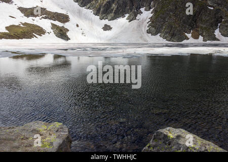 Das Auge Lake im Rila Gebirge in Bulgarien, die von bunten gebrochene Eis bedeckt, felsige Klippen mit Schnee und Vordergrund Felsen mit Moos Stockfoto