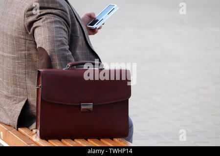 Mann in einem Anzug mit einem Leder Aktentasche auf einer Bank und Sie ihr Smartphone. Konzept für Unternehmer, Beamte, Mitarbeiter Stockfoto