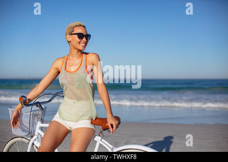 Schöne glückliche Frau in Sonnenbrille sitzt auf Fahrrad am Strand in der Sonne Stockfoto