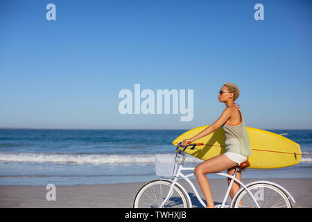 Frau mit Surfboard sitzen auf dem Fahrrad am Strand in der Sonne Stockfoto