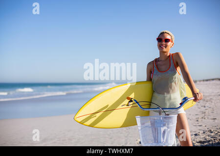 Frau mit Surfboard sitzen auf dem Fahrrad am Strand in der Sonne Stockfoto