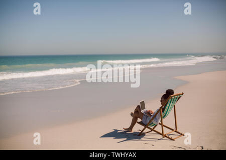 Frau sitzt auf der Liege und mit Laptop am Strand in der Sonne Stockfoto