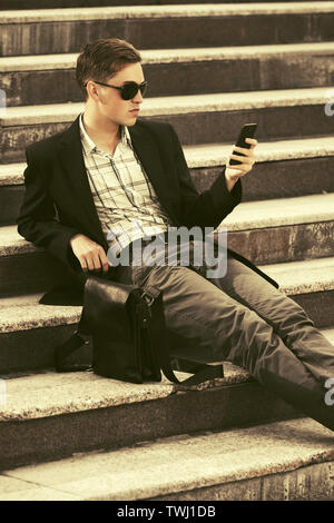 Young Business Mann mit Handy sitzen auf Schritte Stilvolle männliches Modell in Schwarz Blazer und Hose grau Stockfoto