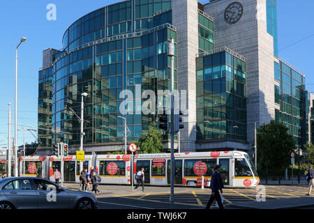 Menschen zu Fuß in einem Dubliner Straße an einem sonnigen Sommerabend mit einer Straßenbahn Luas in Dublin vor ein modernes Gebäude mit einer Glasfassade. Stockfoto