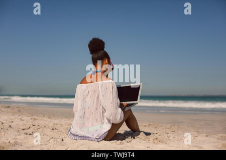 Frau sitzt auf Sand und mit Laptop am Strand in der Sonne Stockfoto