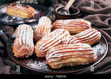 Leckere hausgemachte Eclairs bestreut mit Puderzucker und Schokolade Tropfen auf einem Steingut Teller auf einem dunklen rustikalen alten Tisch, horizontale Ansicht aus Stockfoto