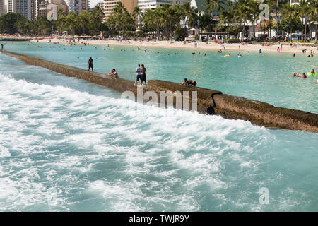 Die ankommenden Wellen schlagen die Ufermauer während Touristen und Einheimische auf der Kuhio Beach Park in Waikiki Beach Walk, Oahu, Hawaii. Stockfoto
