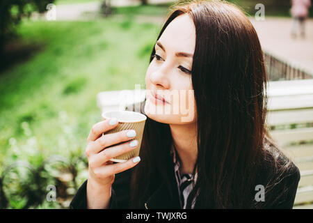 Nett Lächeln, brünett, eine Tasse Kaffee und riechen das Aroma. Charmante business Frau sitzen auf einer Bank in einem Park. Stockfoto