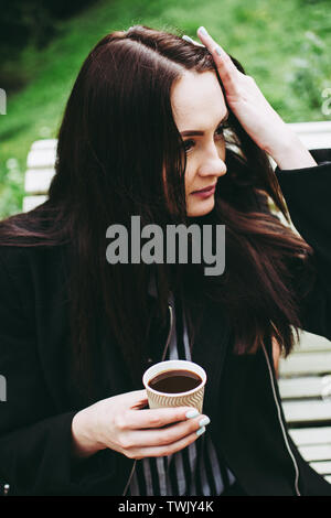 Wunderschöne black-eyed brunette Holding eine Tasse schwarzen Kaffee und Suchen in der Entfernung. Charmante junge Mädchen mit einem schwarzen Mantel und gestreift schwarz a Stockfoto