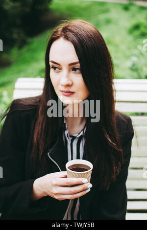 Wunderschöne black-eyed brunette Holding eine Tasse schwarzen Kaffee und Suchen in der Entfernung. Charmante business Frau trug einen schwarzen Mantel und gestreifte Bla Stockfoto