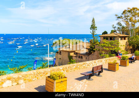 CALELLA DE PALAFRUGELL, SPANIEN - Jul 6, 2019: Touristen sitzen auf der Uferpromenade im malerischen Fischerdorf mit einem kleinen Schloss und Sandstrand mit Cl Stockfoto