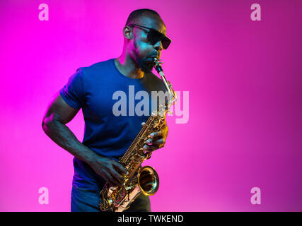 Junge afrikanische-amerikanischen Jazz musiker Saxophon auf Pink Studio Hintergrund in trendigen Neon Licht. Konzept der Musik, Hobby. Freudige attraktiver Kerl improvisieren. Bunte Porträt des Künstlers. Stockfoto