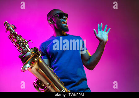 Junge afrikanische-amerikanischen Jazz musiker Saxophon auf Pink Studio Hintergrund in trendigen Neon Licht. Konzept der Musik, Hobby. Freudige attraktiver Kerl improvisieren. Bunte Porträt des Künstlers. Stockfoto
