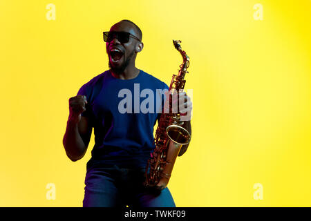 Junge afrikanische-amerikanischen Jazz musiker Saxophon auf Gelb studio Hintergrund in trendigen Neon Licht. Konzept der Musik, Hobby. Freudige attraktiver Kerl improvisieren. Bunte Porträt des Künstlers. Stockfoto