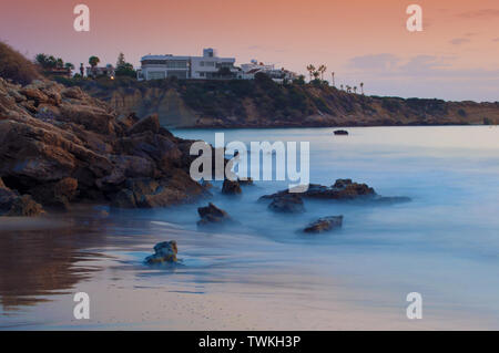 Anzeigen von leeren Sandstrand Coral Bay Beach in der Nähe von Paphos, raucherregelung. Sonnenuntergang, roten Himmel über hellblau Flachwasser, Villen auf der Klippe. Warmen Abend im Herbst. Co Stockfoto