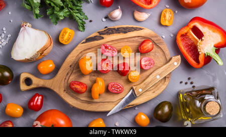 Kirschtomaten in Scheiben geschnitten auf einem Holzbrett unter anderem Gemüse zum Kochen auf grauem Hintergrund. Ansicht von oben, flach Stockfoto