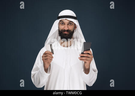 Portrait von arabischen saudischen Geschäftsmann auf dunkelblau studio Hintergrund. Mann mit Smartphone für die Zahlung der Rechnung, Online Shopping oder Wetten. Konzept der Business, Finanzen, Mimik, menschliche Gefühle. Stockfoto