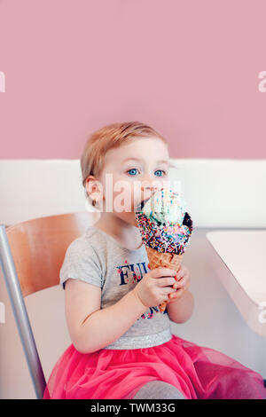 Cute adorable lustig Kaukasischen blond babyl Mädchen mit blauen Augen Essen lecken Eis in große Waffel Kegel mit bunten Streuseln. Happy Chil Stockfoto