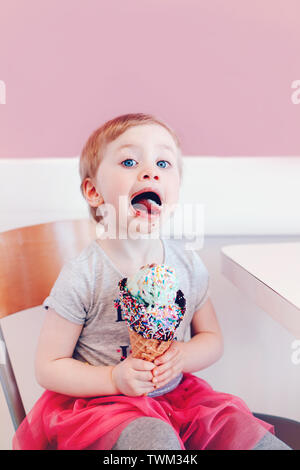 Lustige Kaukasischen blond babyl Mädchen mit blauen Augen Essen lecken Eis in große Waffel Kegel mit Streuseln und zeigt Zunge Süß. Happy chi Stockfoto