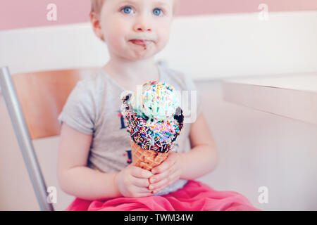 Cute adorable lustig Kaukasischen blond babyl Mädchen mit blauen Augen Essen lecken Eis in große Waffel Kegel mit bunten Streuseln. Happy Chil Stockfoto