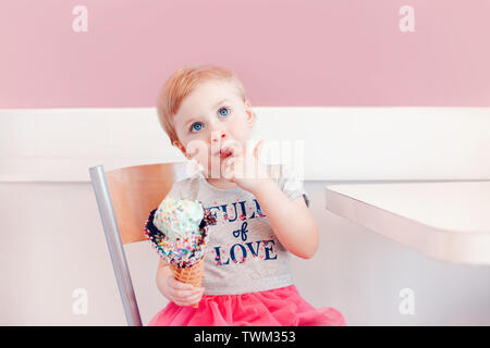 Cute adorable lustig Kaukasischen blond babyl Mädchen mit blauen Augen Eis essen in große Waffel Kegel mit bunten Streuseln. Kid lecken Finger Stockfoto