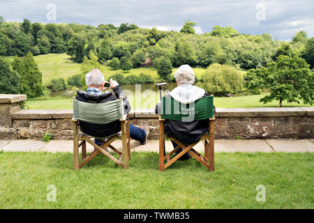 Ein älterer grauhaariger Paar sitzt im Verwaltungsrat Stühle auf dem Rasen an Chartwell Landsitz mit Blick über den See zu den Bäumen Stockfoto