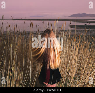 Die Rückseite einer Frau mit langen blonden Haaren Spaziergänge durch die long tall Gräser in Richtung der Großen Salz See bei Sonnenuntergang. Stockfoto