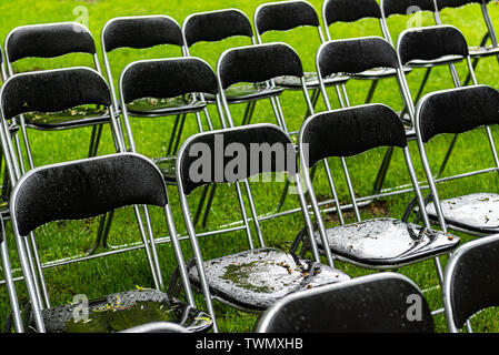 Black Metal Stühle im Freien stehen im Park im Regen. Leere Auditorium, grünes Gras, Bäume und Stühlen mit Wassertropfen. Stockfoto