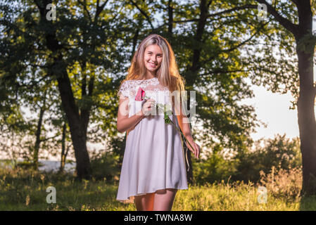 Der lächelnde Modell vor der Kamera. Unschuldig, mit einer Rose in der Hand (ein Symbol der Liebe), mit einem schönen Hintergrund, mit blonden Haaren sanft Tou Stockfoto